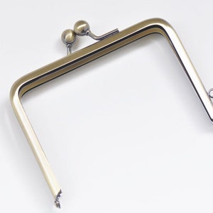 Puede incluir: Un marco de bolso de metal dorado con una parte superior redondeada y dos bolas decorativas. El marco está abierto y listo para ser fijado a un bolso.