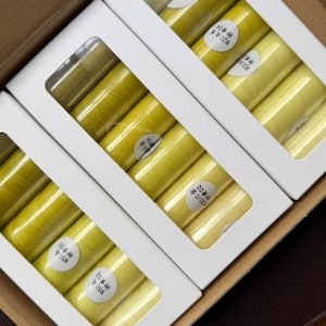 May include: A close-up shot of several boxes of yellow thread spools. Each spool is labeled with a white sticker. The spools are arranged in white boxes with clear windows, and the boxes are inside a larger brown box.