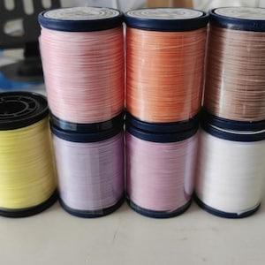 May include: A collection of colorful spools of thread. The spools are in shades of pink, orange, brown, yellow, purple, and white. Each spool has a black base and a dark blue top. The threads are tightly wound, suggesting they are new.