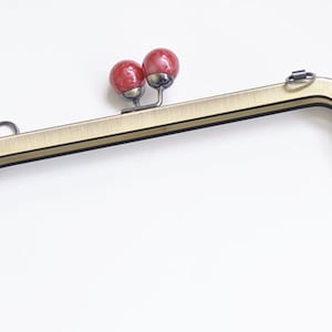 Może przedstawiać: Złoty metalowy stelaż do torebki z dwoma czerwonymi, okrągłymi ozdobami. Stelaż jest przeznaczony do użytku w torebce lub kopertówce.