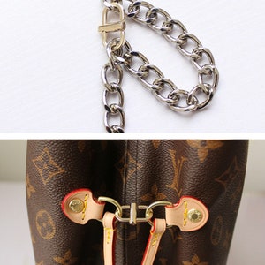 May include: Close-up of a brown leather handbag with a gold metal chain strap. The bag has a brown and beige monogram pattern and a gold metal clasp.