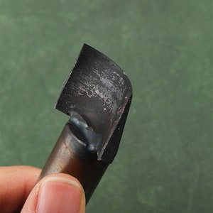 May include: A close-up of a black metal woodworking tool with a curved blade. The tool is held in a hand.