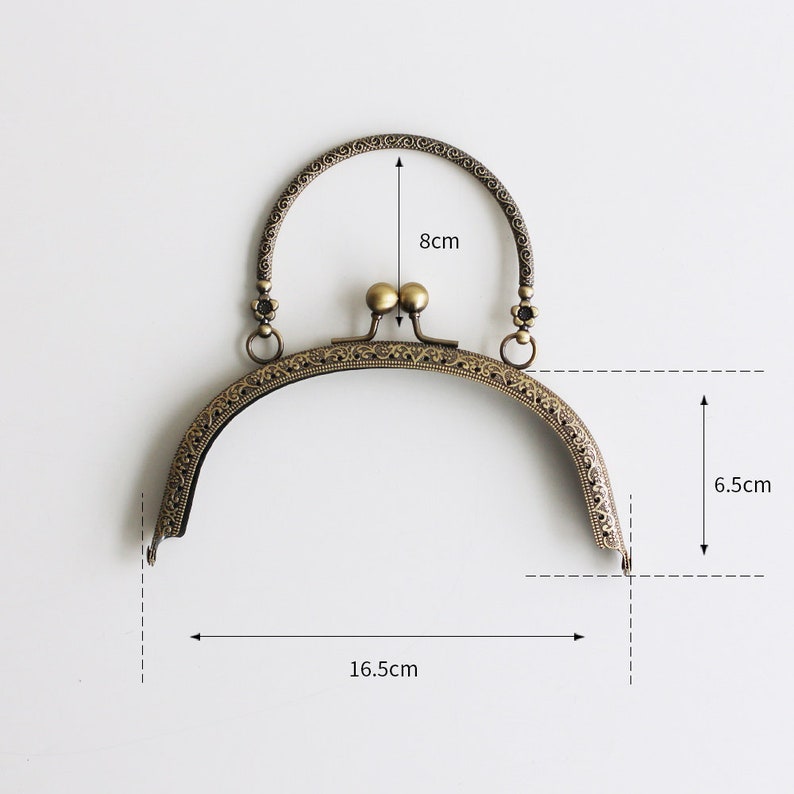 May include: A gold-toned metal purse frame with an ornate design. The frame measures 6.5 inches tall, 16.5 inches wide, and has a handle that is 8 inches long.