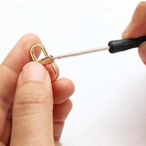 May include: A person uses a small screwdriver to tighten a gold-colored metal clasp. The clasp has two loops.