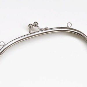 May include: A silver-colored metal purse frame with a curved, arched design. The frame features two small ball-shaped accents at the top center and small loops on each side for attaching to a purse body. The frame is designed for crafting.