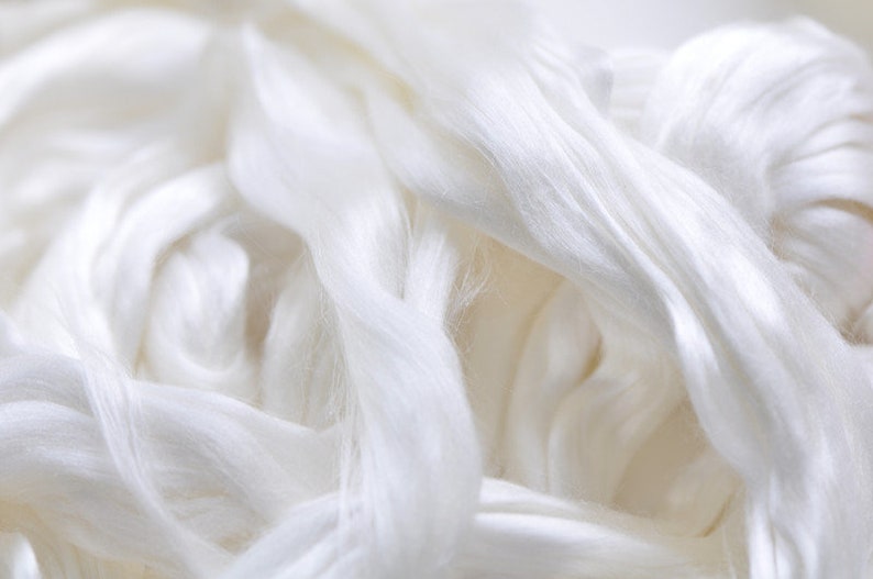 May include: Close-up of a pile of soft, white silk fibers. The fibers are tightly packed and have a smooth, flowing texture. The image highlights the natural sheen and delicate appearance of the silk, suggesting its use in textiles or crafting.
