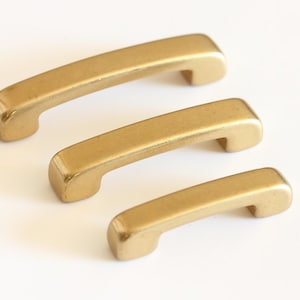 May include: Three gold-toned metal cabinet pulls in different sizes. The pulls are shaped like a rounded rectangle with a curved top.