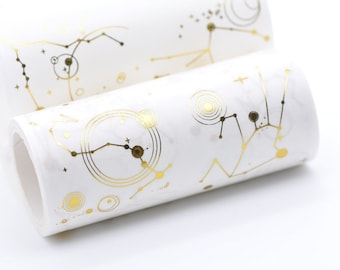 May include: A roll of white washi tape with gold foil constellations and circles. The tape is rolled up and the end is visible.