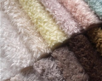 May include: Close-up of a variety of plush, textured fabrics in a range of colors. The colors include cream, pale yellow, pink, brown, and teal. The fabrics have a soft, fluffy appearance, suggesting they are made of a synthetic material.