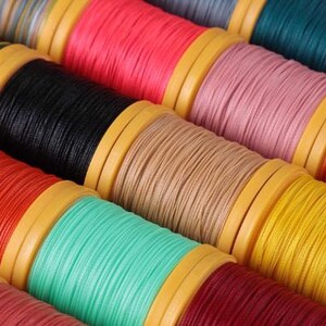 May include: Close-up of colorful spools of thread. The spools are arranged in rows, showcasing a variety of colors including red, pink, black, tan, teal, yellow, and orange. The thread is tightly wound on yellow plastic spools.