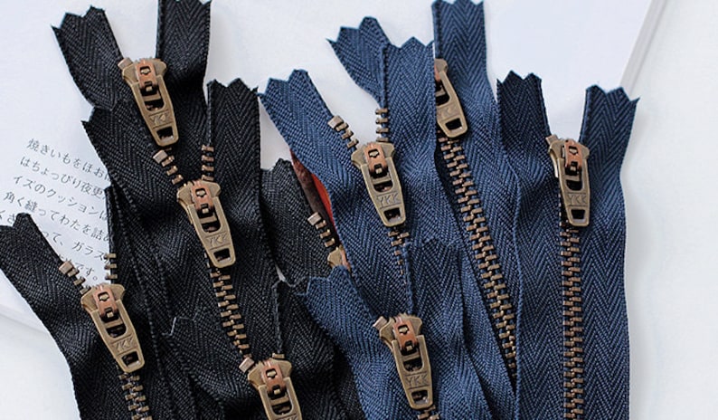 K&ouml;nnte beinhalten: Nahaufnahme einer Gruppe von schwarzen und blauen Rei&szlig;verschl&uuml;ssen mit Messing-Zierb&auml;ndern. Die Rei&szlig;verschl&uuml;sse sind in einem zuf&auml;lligen Muster angeordnet und alle ge&ouml;ffnet.