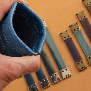 May include: A blue leather coin purse with a zipper closure. The purse is being held in a hand. There are five metal clasps with a blue and silver finish on a brown surface.