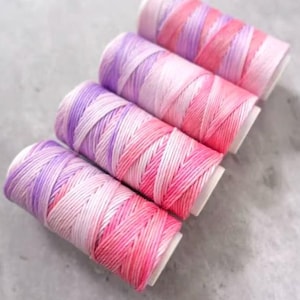 May include: Four spools of colorful thread in shades of pink, purple, and white. The thread is tightly wound around white cardboard spools. The colors blend together, creating a gradient effect. Ideal for crafting and sewing projects.