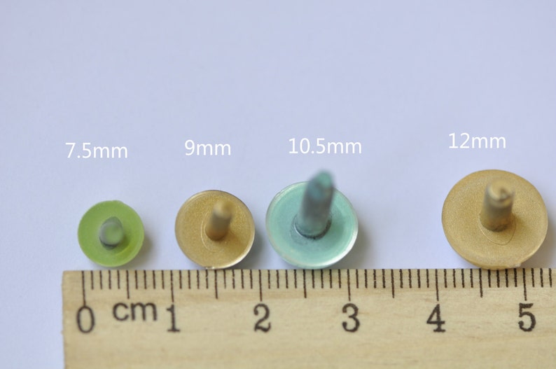 May include: Four metal buttons in different sizes, 7.5mm, 9mm, 10.5mm, and 12mm, with a ruler for scale. The buttons are gold, green, and blue.