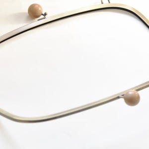 May include: A metal purse frame with a rectangular shape and rounded corners. The frame has a light gold finish and features two small, round, wooden-colored knobs. The frame is designed to be used for making handbags or purses.