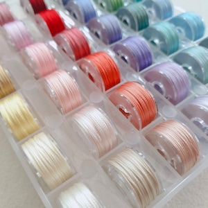 Könnte beinhalten: Eine transparente Plastik-Aufbewahrungsbox gefüllt mit Garnrollen in verschiedenen Farben, darunter Rot, Rosa, Orange, Gelb, Grün, Blau und Lila.