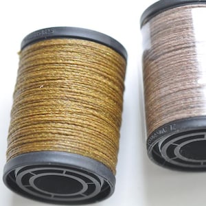 Può includere: Due rocchetti di filo. Uno è di colore oro metallizzato e l'altro marrone. Entrambe le bobine hanno una base e una parte superiore nere. Il filo è avvolto strettamente attorno alla bobina.