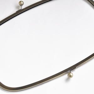 May include: A bronze-colored purse frame with a rounded rectangular shape. The frame has a ball clasp at each end and is designed to be sewn into a bag. The metal has a slightly aged appearance.