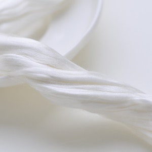 May include: Close-up of a bundle of white silk fibers, appearing soft and flowing. The fibers are arranged in a twisted, rope-like formation, suggesting a natural textile material. The background is a white, shallow dish.