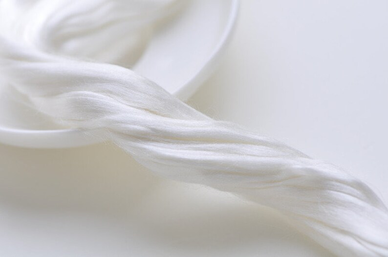 May include: Close-up of a bundle of white silk fibers, appearing soft and flowing. The fibers are arranged in a twisted, rope-like formation, suggesting a natural textile material. The background is a white, shallow dish.
