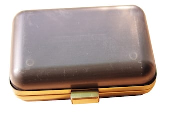 Può includere: Una piccola pochette rettangolare con una cornice e una chiusura in metallo dorato. La pochette è realizzata in materiale grigio scuro con una finitura liscia e leggermente testurizzata.