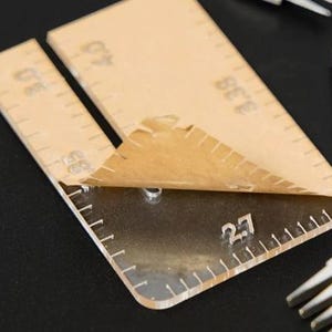 Op de afbeelding: Een doorzichtig acryl gereedschap voor leerbewerking met gegraveerde afstandsmarkeringen waaronder 2.7, 3.0, 3.38 en 4.0. Een deel van de bruine beschermende papieren laag is gedeeltelijk losgetrokken van het oppervlak van het gereedschap.