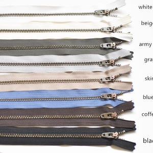 May include: An assortment of zippers in shades such as white, beige, army green, grey, skin tone, blue, coffee, and black. Each zipper features a metal pull and teeth, with colour labels alongside.