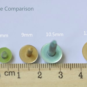 May include: Size comparison of four round, flat-topped, decorative metal tacks with a pointed tip. The tacks are 7.5mm, 9mm, 10.5mm, and 12mm in diameter. The tacks are green, gold, blue, and gold in color. The tacks are lined up on a wooden ruler with centimeter markings.