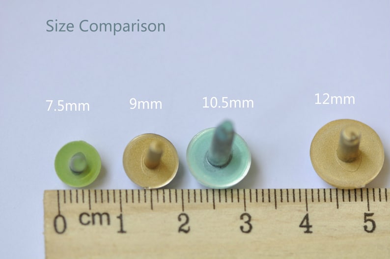 May include: Size comparison of four round, flat-topped, decorative metal tacks with a pointed tip. The tacks are 7.5mm, 9mm, 10.5mm, and 12mm in diameter. The tacks are green, gold, blue, and gold in color. The tacks are lined up on a wooden ruler with centimeter markings.