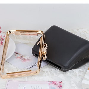 Pode incluir: Uma moldura de metal dourado para uma bolsa de mão com um painel de acrílico transparente. A moldura está aberta e a bolsa está fechada, mostrando um exterior de tecido preto.