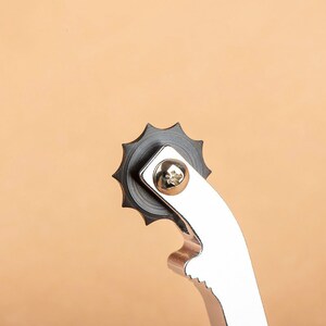 Puede incluir: Primer plano de una herramienta de metal plateada con una rueda negra en forma de estrella. La rueda tiene un tornillo en el centro.
