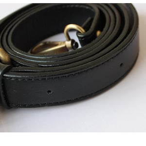 May include: A black leather strap with a gold-colored metal clasp. The strap is rolled up, showing its smooth texture and stitching. The clasp is attached to the strap with a small, black piece.
