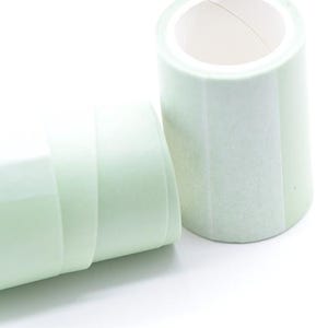 Könnte beinhalten: Zwei Rollen hellgrünes Washi-Tape vor weißem Hintergrund. Eine Rolle steht aufrecht und zeigt den inneren Kern, während die andere auf der Seite liegt. Das Klebeband hat ein subtiles, strukturiertes Aussehen.