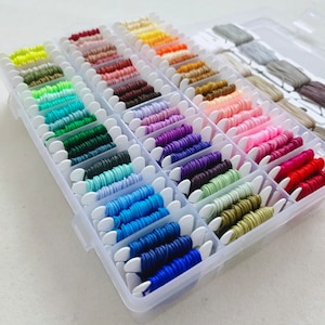 May include: A clear plastic storage box filled with rows of colorful embroidery floss spools. The spools are organized by color, with each row containing a different shade.