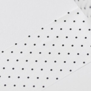 May include: White washi tape with a repeating pattern of small black polka dots. The tape is partially unrolled, revealing the roll's edge. The background is a plain white surface.