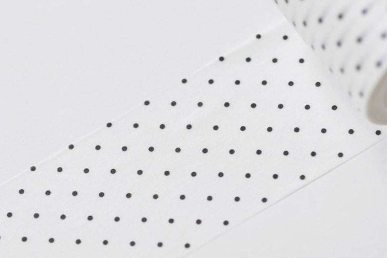 May include: White washi tape with a repeating pattern of small black polka dots. The tape is partially unrolled, revealing the roll's edge. The background is a plain white surface.