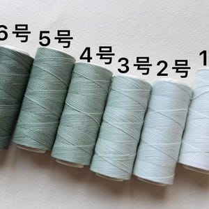May include: Six spools of thread in varying shades of green and blue. The spools are labeled with Japanese numerals from 1 to 6. The thread is tightly wound on the spools, creating a textured appearance. The colors range from a deep green to a light blue.