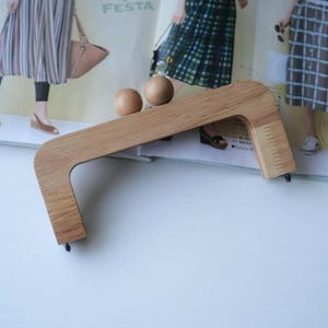 Könnte beinhalten: Ein hellbrauner Holz-Taschengriff mit geschwungenem Design und zwei kleinen Holzkugeln. Der Griff ist abgewinkelt und hat eine rechteckige Form mit abgerundeten Ecken. Der Hintergrund zeigt eine Zeitschrift mit Bildern von Personen.