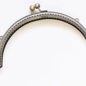 May include: A vintage-style, arched purse frame in an antique brass finish. The metal frame features intricate embossed detailing along the edges and a clasp with two small, round knobs. Perfect for crafting handmade bags and clutches.