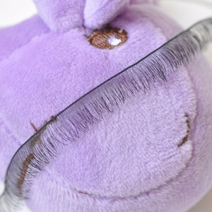 Puede incluir: Un juguete de peluche morado con pestañas negras pegadas en la parte superior. El juguete tiene una nariz y una boca bordadas en marrón.