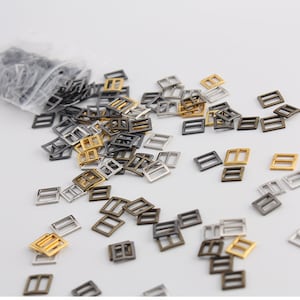 Op de afbeelding: Een zak met verschillende metalen schuifgespen in goud, zilver en brons. De gespen zijn vierkant met een rechthoekige opening in het midden.