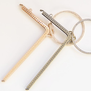 Puede incluir: Dos marcos de bolso de metal, uno dorado y otro de latón antiguo, con agujeros decorativos y un borde redondeado. Los marcos están diseñados para ser utilizados para crear bolsos o bolsas.