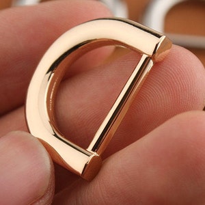 Puede incluir: Un anillo en D de metal dorado con un acabado liso y pulido. El anillo tiene un diámetro de aproximadamente 2,5 cm.