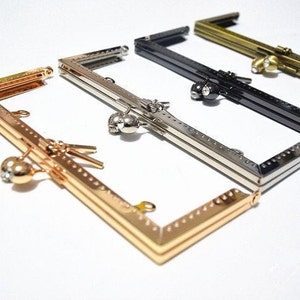 May include: Four different metal purse frames, each with a different finish: gold, silver, black, and bronze. The frames are all rectangular and have a decorative design. Each frame has a small, round, decorative knob on the side.