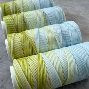 May include: Four spools of colorful thread. The thread transitions from yellow-green to light blue. The spools are white and arranged in a diagonal line. The thread is tightly wound, and the colors blend together.