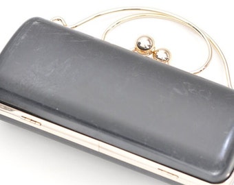 Può includere: Una pochette nera con un manico superiore in metallo dorato e due sfere in metallo dorato. La pochette ha una forma rettangolare e una finitura liscia e lucida.