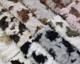 May include: Close-up of a pile of faux fur fabric in various colors. The fabric has a short, textured pile and features a pattern of brown, black, white, and gray patches. The overall effect is soft and plush.