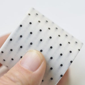 May include: A roll of white washi tape with a repeating pattern of small black polka dots. The tape is held in a person's hand, showcasing the design and texture. Ideal for crafts and decorative projects.