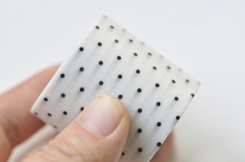 May include: A roll of white washi tape with a repeating pattern of small black polka dots. The tape is held in a person's hand, showcasing the design and texture. Ideal for crafts and decorative projects.