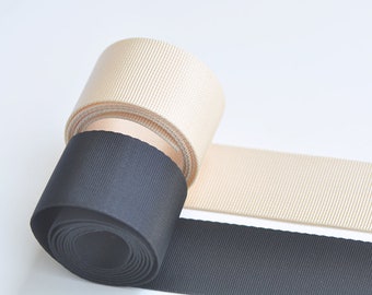 May include: Two rolls of ribbon, one black and one cream, are shown on a white surface. The black ribbon is rolled up and the cream ribbon is partially unrolled.